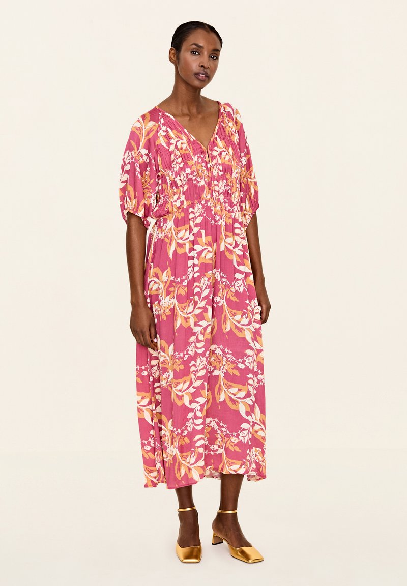 Rochie maxi floral roz cu mâneci bufante, decolteu în V, talie strânsă și pantofi cu toc bloc aurii. Material neted cu un model vibrant.