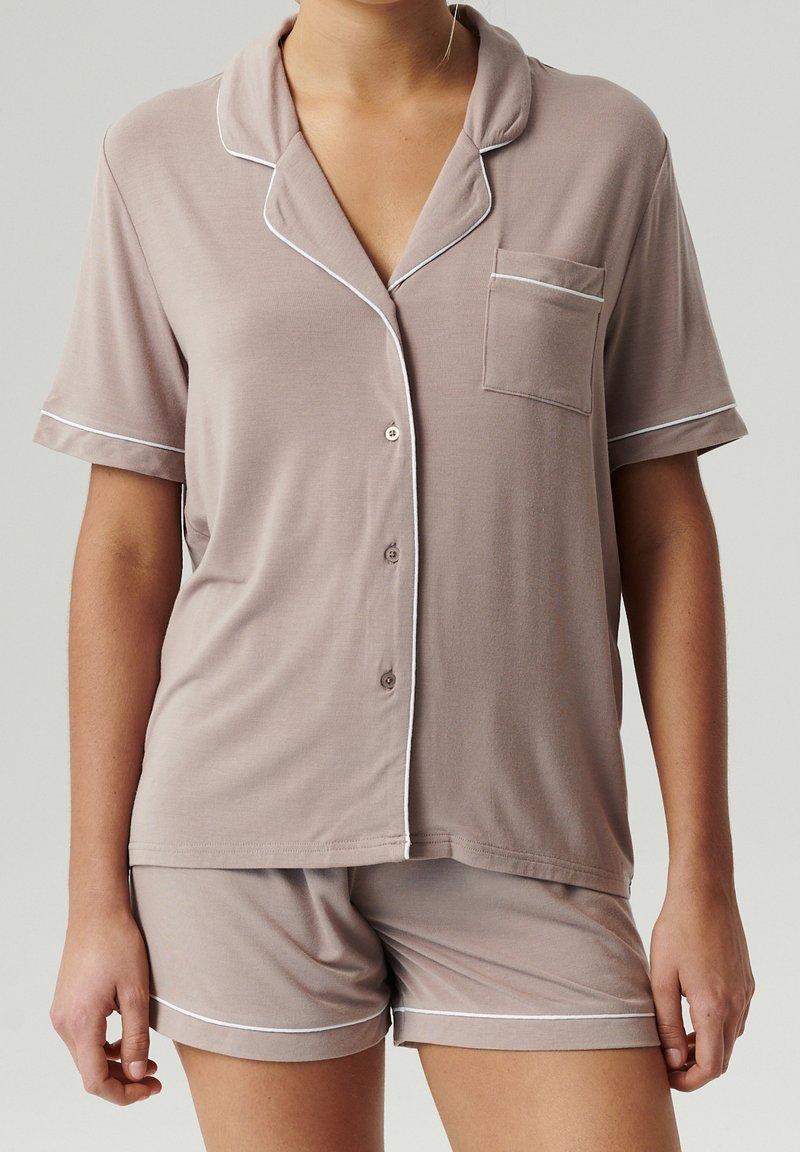 Kobieta w taupe'owej koszuli z krótkim rękawem zapinanej na guziki i pasujących szortach z białym wykończeniem, stojąca na jednolitym tle.