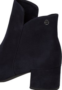 Bottine en suede bleu marine avec un petit talon carré, présentant un design découpé en haut et un accent métallique rond sur le côté.