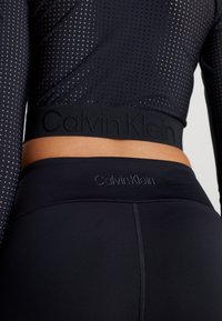 Top corto sportivo nero con design perforato e fascia con logo Calvin Klein, abbinato a leggings neri a vita alta con logo in rilievo.