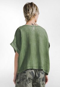 Person mit kurzen blonden Haaren, die ein locker sitzendes grünes Oberteil und gemusterte Hosen trägt, mit einem Schmetterlingstattoo auf dem oberen Rücken und einem kleinen Handtattoo.