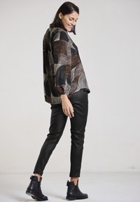 Smiling woman in black leather pants, black ankle boots, and a brown, black, and beige patterned long-sleeve blouse standing against plain background.