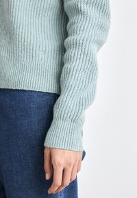 Helles blaues geripptes Strickpullover mit langen Ärmeln, der eine strukturierte Oberfläche aufweist. Getragen mit dunkelblauen Jeanshosen.