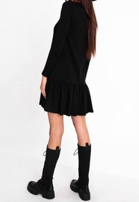 Schwarzes langärmliges Kleid mit ausgestelltem Saum, aus weichem Stoff. Getragen mit gerippten schwarzen knielangen Socken und klobigen schwarzen Stiefeln.