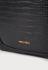 Borsa nera con texture al coccodrillo, caratterizzata da un logo dorato impresso sulla parte frontale. Bordo liscio e forma strutturata.