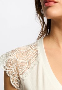 Top bianco con dettagli in pizzo intricati sulle spalle, con una morbida texture a costine e un design a scollo a V.