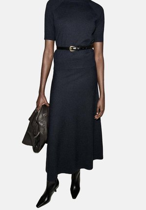 Vestido marino de tela de punto con mangas cortas, acentuado por un cinturón negro. La modelo sostiene una chaqueta de cuero oscura y lleva botas negras.