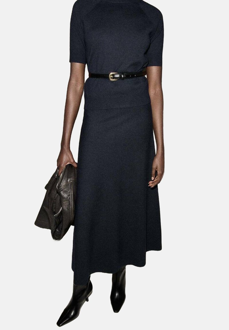 Vestido marino de tela de punto con mangas cortas, acentuado por un cinturón negro. La modelo sostiene una chaqueta de cuero oscura y lleva botas negras.