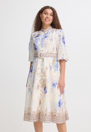 Femme aux cheveux bouclés portant une robe midi crème avec imprimé floral bleu, manches bouffantes, col haut et ceinture large avec boucle circulaire.