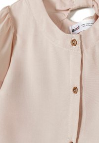 Blouse rose clair pour tout-petit avec des manches bouffantes, un col rond et deux petits boutons marron sur le devant.