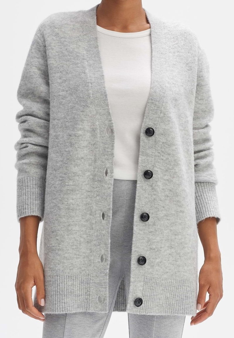Personne portant un long cardigan gris clair avec des boutons noirs sur une chemise blanche et un pantalon gris assorti, debout les bras détendus.