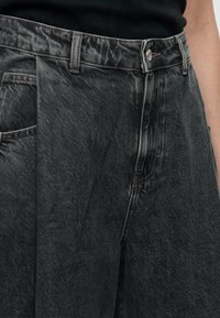 Gros plan d'une personne portant un jean noir délavé taille haute avec un bouton visible et des passants pour ceinture.