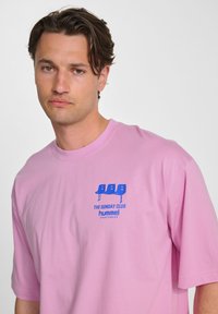 Rosa bomulls t-shirt med en avslappnad passform, som har en blå grafisk design på bröstet som inkluderar stiliserade stolar och text.