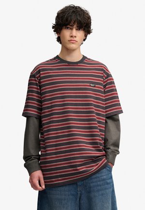 Junger Mann mit lockigem Haar, der ein mehrlagiges rot-schwarz gestreiftes kurzärmeliges T-Shirt über einem grauen Langarmshirt und blauen Jeans trägt, steht.