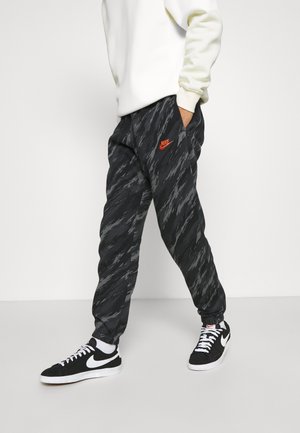 Personne portant un jogging Nike noir et gris à motifs avec un logo orange, des baskets Nike noires et blanches, et un sweat-shirt blanc.