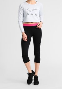 Sweat-shirt court gris clair avec le texte "ENTRAÎNEMENT", associé à un legging capri noir avec une ceinture rose. Chaussures de sport noires.