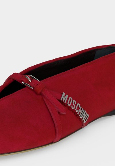 Chaussure plate en daim rouge avec une bride décorative et un logo métallique argenté "MOSCHINO" sur le côté.
