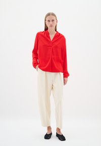 Blusa rossa a maniche lunghe con scollatura a V e leggero lucido, abbinata a pantaloni beige a vestibilità ampia. Ai piedi, scarpe slip-on nere con texture completano il look.