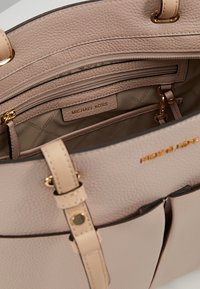 Jasnoróżowa skórzana torebka o teksturowanym wykończeniu, złotych akcesoriach i wielu wewnętrznych przegrodach z materiałową podszewką i metką z logo.