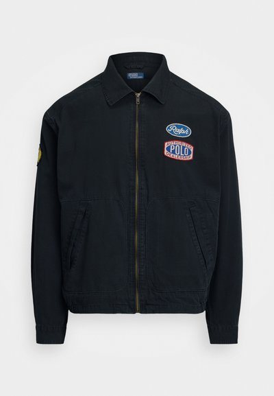 Must must, mis pehmeast kangast, esifliis, kahe küljetasku ja mitme tikandiga plaastriga rinnal ja varrukal.