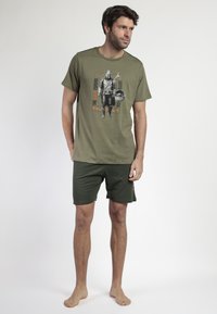 Hombre vestido con pantalones cortos verde oliva y una camiseta con un gráfico de Mandalorian y Grogu, de pie descalzo frente a un fondo blanco liso.
