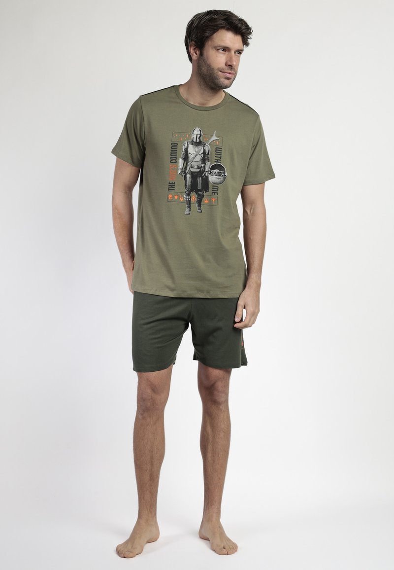 Hombre vestido con pantalones cortos verde oliva y una camiseta con un gráfico de Mandalorian y Grogu, de pie descalzo frente a un fondo blanco liso.