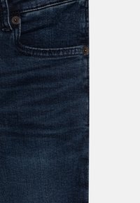 Granatowe dżinsy z denimu w klasycznym kroju z pięcioma kieszeniami. Materiał ma lekko zużyty wygląd z detalami szwów i metalowymi ozdobami.