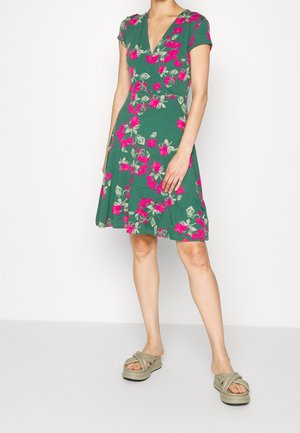 Femme portant une robe verte longueur genou avec un imprimé floral rose et des sandales compensées beiges, debout devant un fond clair uni.