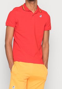 Czerwona koszulka polo z kołnierzykiem w żółte paski, krótkim rękawem i logo K-way na piersi. Noszona z żółtymi szortami. Gładka tekstura.