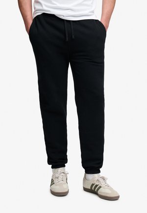 Person iført sorte joggingbukser med elastiske manchetter, hvide sokker og beige sneakers med mørkegrønne striber, hænderne i lommerne.