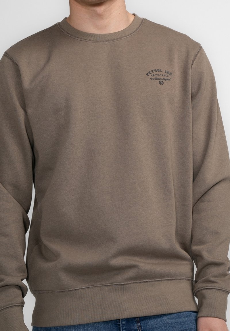 Homme portant un sweat-shirt marron à col rond avec le texte « Petrol Indigo Arctic Race Ice Riders Legend » sur la poitrine gauche, associé à un jean bleu.
