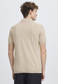 Cămașă polo bej cu mâneci scurte, fabricată din material tricotat, având guler clasic și mâneci cu model în relief, cu un finisaj texturat simplu.