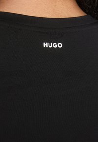 Black T-shirt featuring a slim fit design, made of smooth fabric with a rounded neckline. White "HUGO" logo printed on the back near the collar.