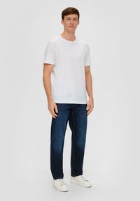 Weißes T-Shirt mit kurzen Ärmeln und Rundhalsausschnitt, kombiniert mit dunkelblauen Jeans und weißen Turnschuhen, vor einem hellgrauen Hintergrund stehend.