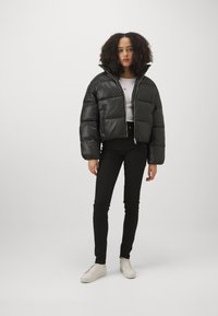 Svart cropped pufferjakke med en glansfull overflate, med høy krage og glidelås foran, kombinert med svarte skinny jeans og hvite sneakers.