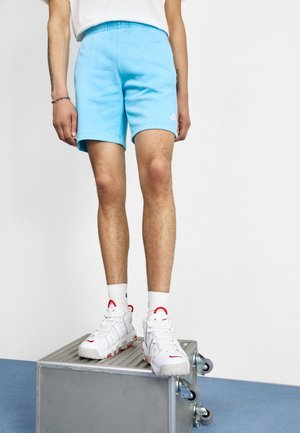 Hellblaue Baumwollshorts mit elastischem Bund, knielang, mit kleinem weißem Logo. Kombiniert mit weißen Sneakern mit roten Akzenten.