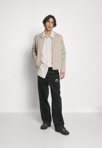 Beige lightweight jacket, collared shirt in light fabric, black pants with Nike logo, and dark sneakers with textured detailing.