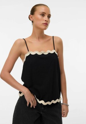 Kvinne iført en svart ermeløs topp med hvit bølget kant og svarte bukser, stående med én hånd på hoften, hvit bakgrunn.