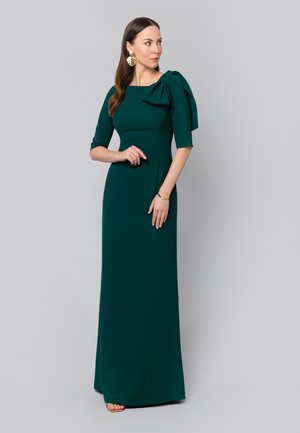 Kobieta w ciemnozielonej, długiej do ziemi sukni z rękawami do łokci i dużą kokardą na lewym ramieniu, nosząca kolczyki w kształcie liści.