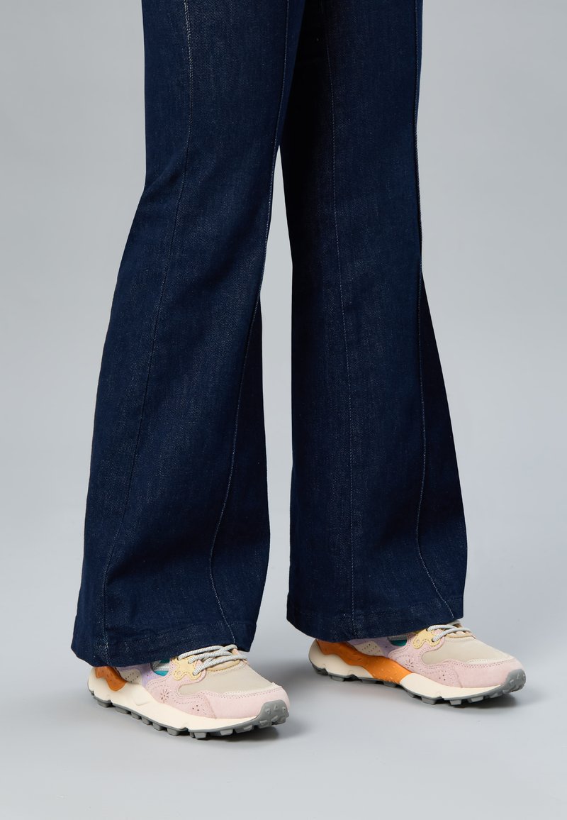 Person trägt dunkle, weit geschnittene Jeans und mehrfarbige Sneakers in Rosa-, Orange- und Beigetönen auf einem hellgrauen Boden.