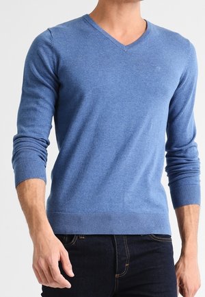 Homme portant un pull bleu ajusté à col en V avec poignets et ourlet côtelés, associé à un jean bleu foncé.