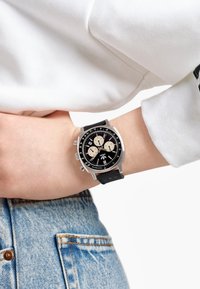 Svart kronografklocka med silverdetaljer och tre underskivor, med ett texturerat svart band, visas på en handled med en vit ärm.