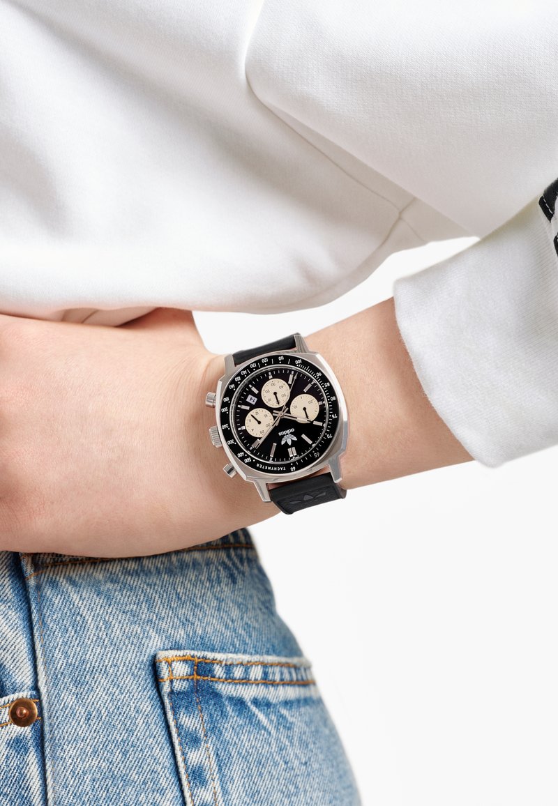 Svart kronografklocka med silverdetaljer och tre underskivor, med ett texturerat svart band, visas på en handled med en vit ärm.