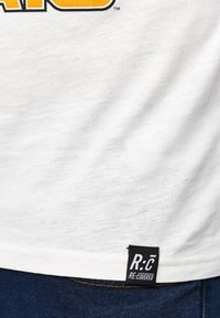 Weißes Baumwoll-T-Shirt mit einem gelben Grafikdruck auf der Vorderseite. Verfügt über ein schwarzes Gewebeschild am Saum mit dem Text "R:C RE:COVERED".