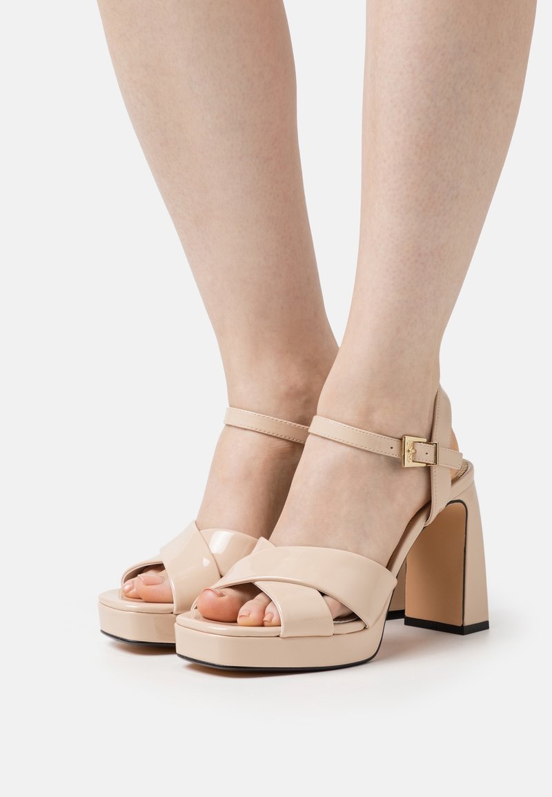 Personne portant des sandales à talons hauts en cuir verni beige, avec brides à la cheville et talons blocs, debout sur un fond clair.