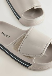 Helle beige Rutschsandalen mit verstellbarem Riemen. Texturiertes Stoffdetail und schwarze Gummisohle mit weißen Streifen. Markenname: NEXT.