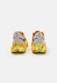Lichtgrijze hardloopschoenen met een gestructureerde bovenkant, oranje accenten en een kleurrijke, sculpted zool in gele en oranje gradaties.