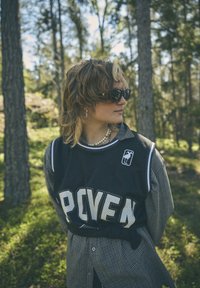 Fekete hálós kosárlabda mez, nagy fehér betűkkel "PROVEN" felirattal, kockás gombos inggel párosítva. Kiegészítők között napszemüveg és gyöngy nyaklánc található.