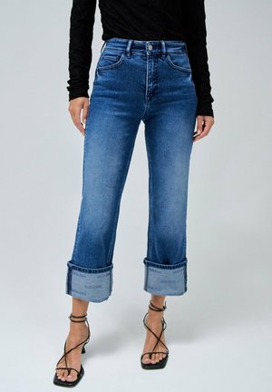 Vrouw draagt hooggetailleerde blauwe jeans met brede omgeslagen boorden en zwarte sandalen met hak en bandjes, gecombineerd met een zwarte top met lange mouwen.