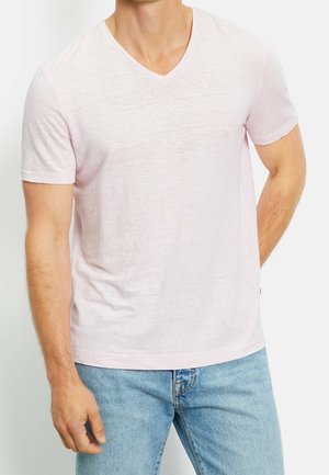 Mann trägt ein hellrosa T-Shirt mit V-Ausschnitt und kurzen Ärmeln sowie hellblaue Jeans, steht mit einer Hand hinter dem Rücken vor weißem Hintergrund.